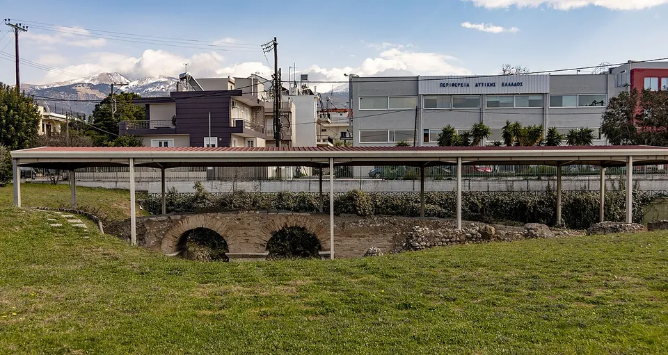 A long open-sided metal shelter over a sunken lawn where two low arches show, with a car park, modern buildings and snow-capped mountains behind.