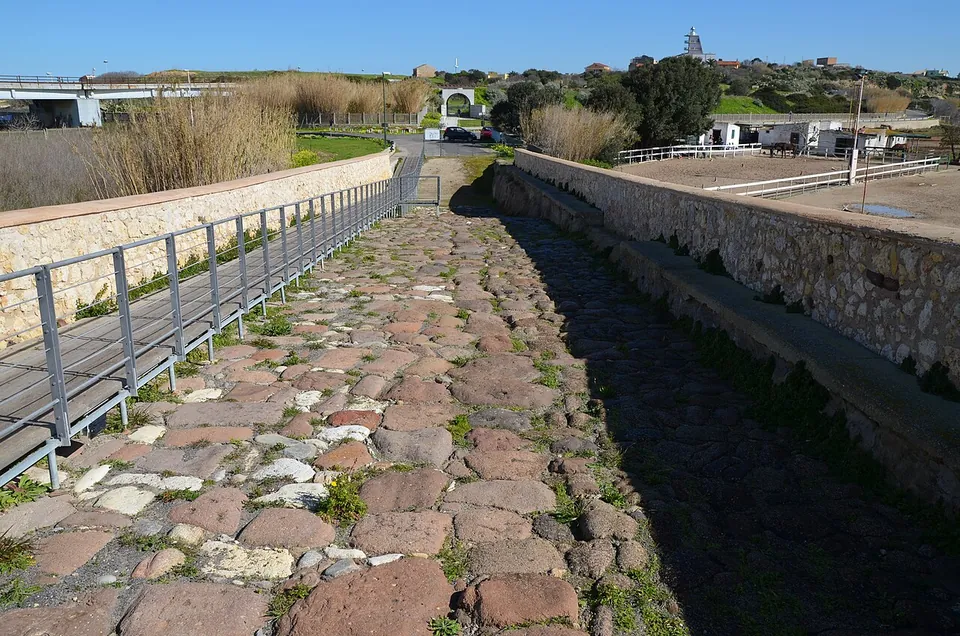 The top of the bridge, its roadway of large rounded paving stones in red and grey running between stone parapets, with a raised walkway of timber boards behind metal railings along the left side.