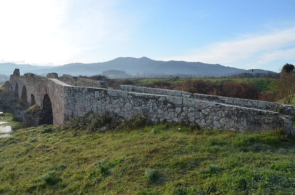 One end of the bridge, where its arches give way to a long stretch of solid masonry running across a grassy bank, with a range of hills under a blue sky behind.