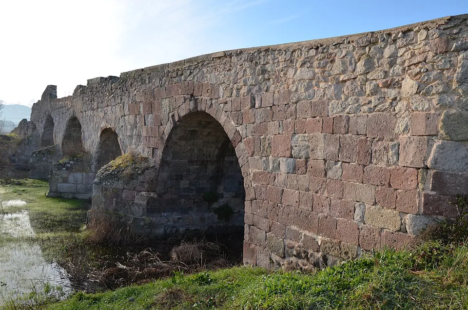 The side of the bridge at close range, a wall of squared reddish and grey stone blocks laid in courses over a round arch, with a pier running down into shallow water.