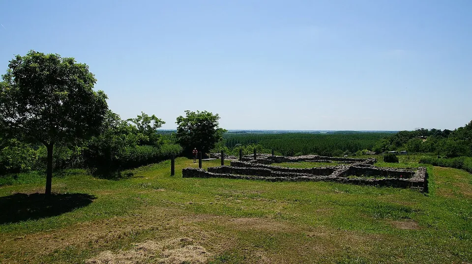 Low stone foundations of a building with several rooms on a mown hilltop, trees on the left and a wide flat plain stretching to the horizon behind.
