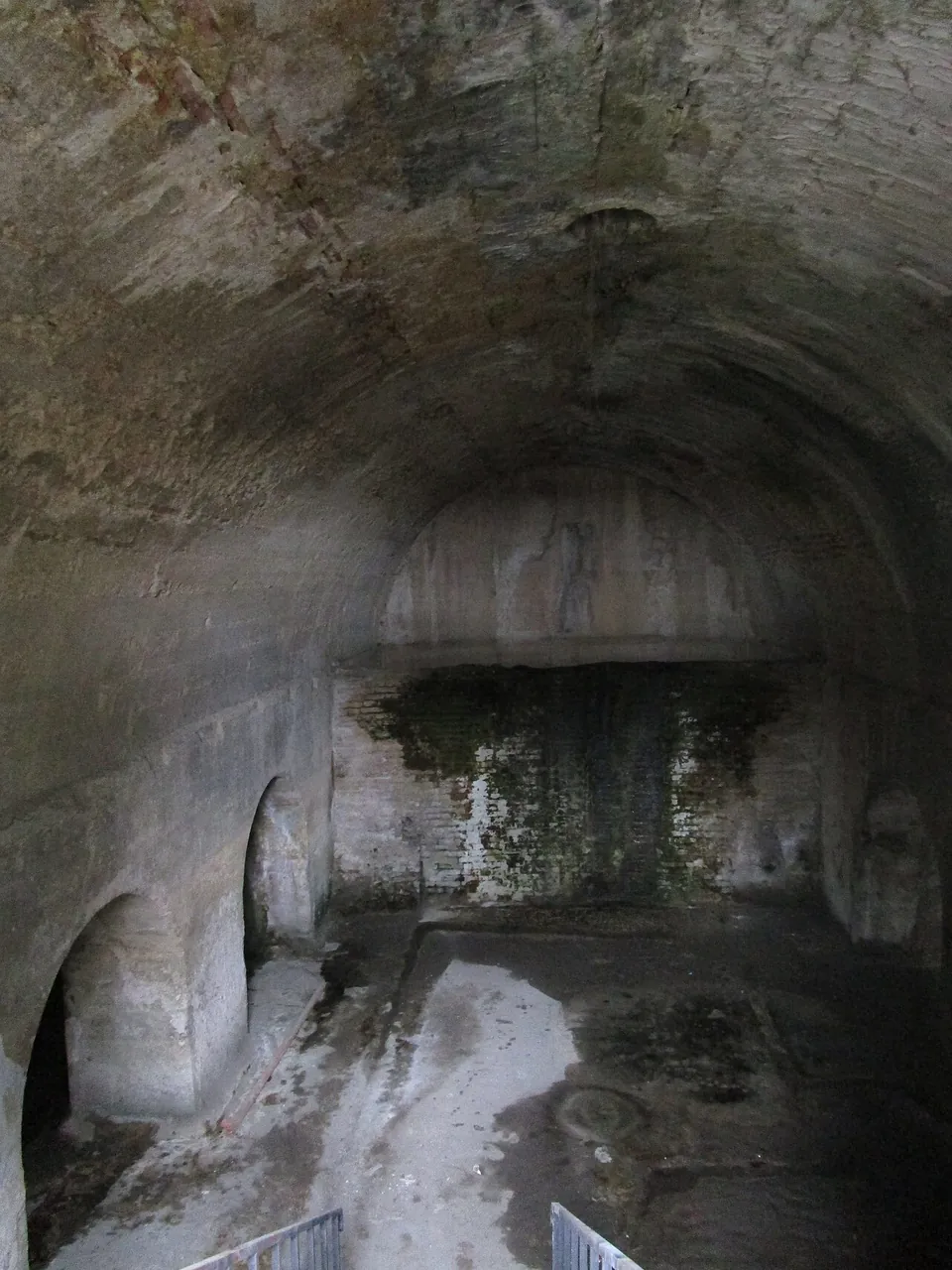 The inside of a long underground chamber with a concrete barrel vault, arched openings along one side and a pale, worn floor.