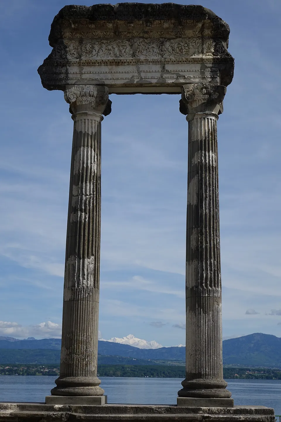 Two tall fluted columns with carved capitals carrying a heavy stone beam decorated with a band of scrolling foliage, against a blue sky, with the lake and a range of mountains with a snow-covered peak far behind.