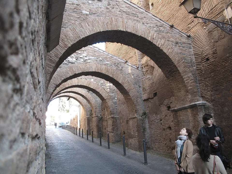 A narrow paved lane running under a long series of brick arches that span it from wall to wall, with tall brick walls on both sides and a few people standing in the foreground.
