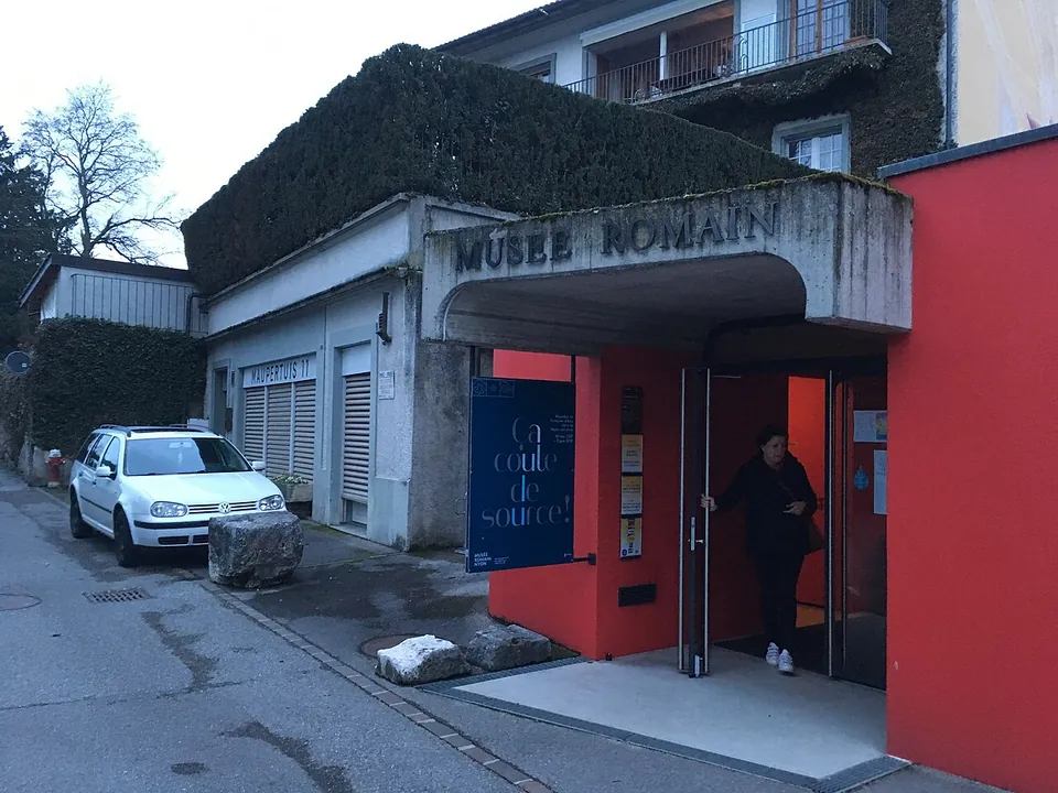 A small entrance with a concrete canopy lettered "Musée romain" and glass doors set in a red wall, on a quiet street with a parked car.