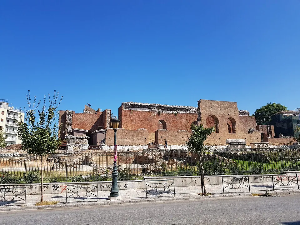 The high brick walls of a ruined stage building with arched openings, seen from a city street through railings, with apartment blocks on either side.