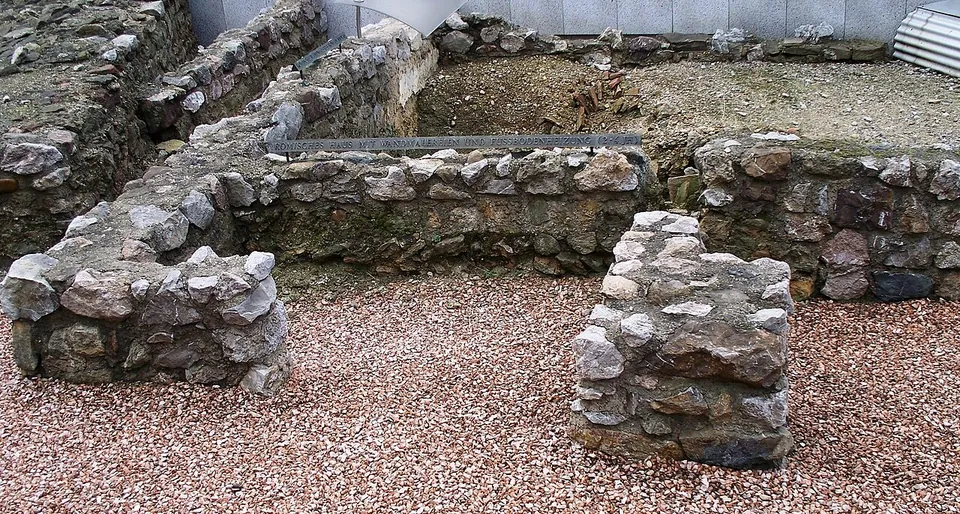 Low walls of rough grey and brown stones, a few courses high, enclosing small rooms floored with reddish gravel, with a metal label strip along one wall and a modern pale wall behind.