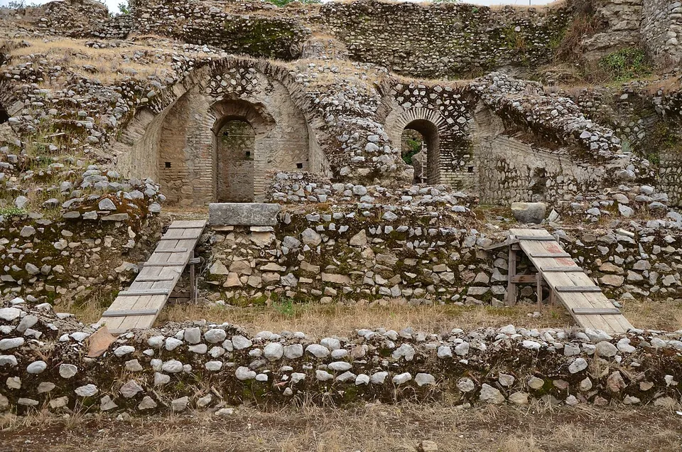 Ruined walls of rubble and brick with two round-arched openings, rising in steps above a low stone wall, with wooden steps leading up between them.