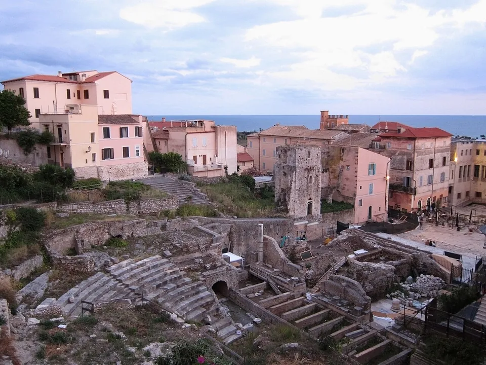 The curved stone seating of a ruined theatre seen from above, cut into by excavation trenches and walkways, with pink and ochre houses of the old town around it and the sea on the horizon.