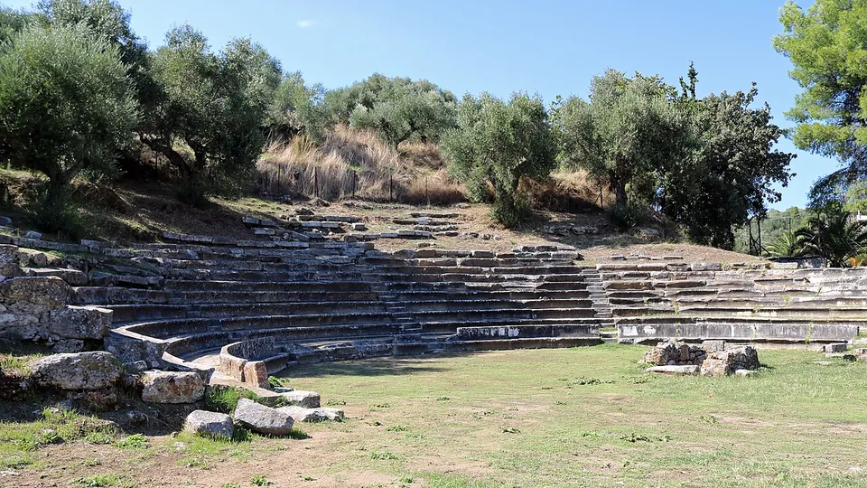 The curved rows of a small stone theatre set into a slope under olive trees, with a grassy orchestra in front.