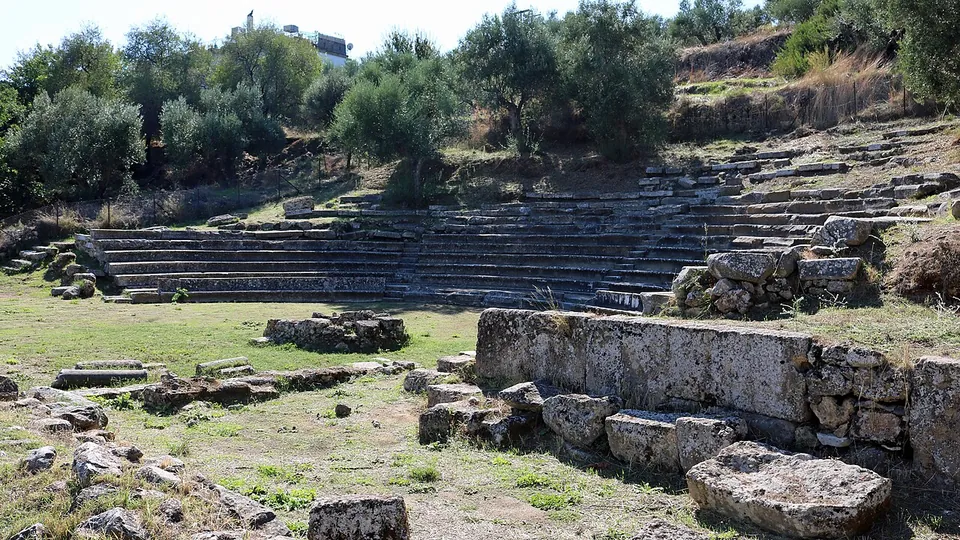 Rows of stone seats in a hollow below olive trees, seen across a grassy orchestra past the squared blocks of a wall in the foreground.