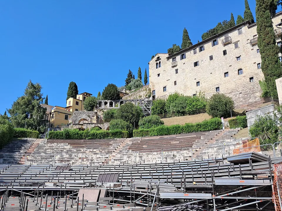 Stone tiers of seating rising up a hillside under a blue sky, their lower part covered by rows of temporary seating and scaffolding, with a long, many-windowed former convent and cypresses above.