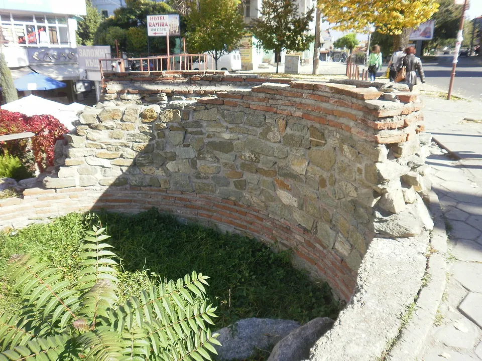 A curved wall of stone with bands of brick enclosing a grassy hollow, beside a pavement where people walk past shops and a hotel sign.