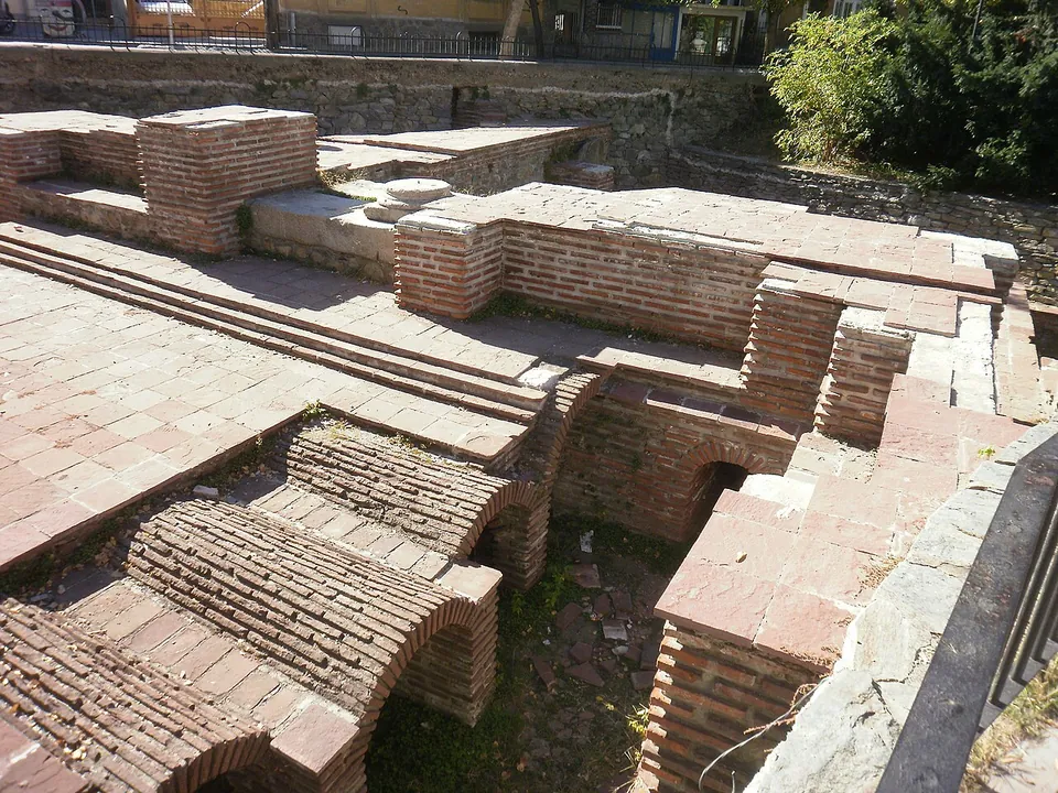 Excavated rooms of brick, with rows of small brick vaults and arched openings below floor level and paved brick surfaces above, enclosed by a railing in a town street.