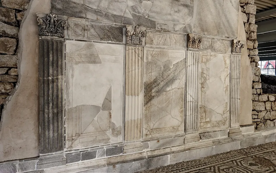 A wall faced with cracked white marble panels framed by fluted pilasters with carved Corinthian capitals, set against rough stonework, with the edge of a mosaic floor in the foreground.