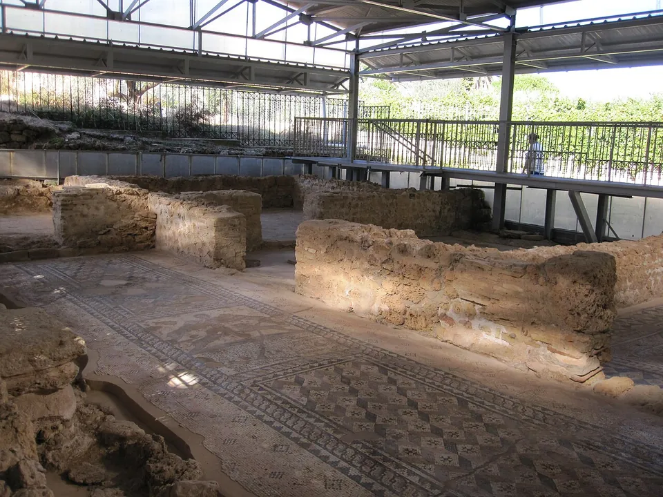 Low ruined walls of rough stone dividing rooms floored with patterned mosaics, under a modern metal roof with a raised walkway and railings along one side.