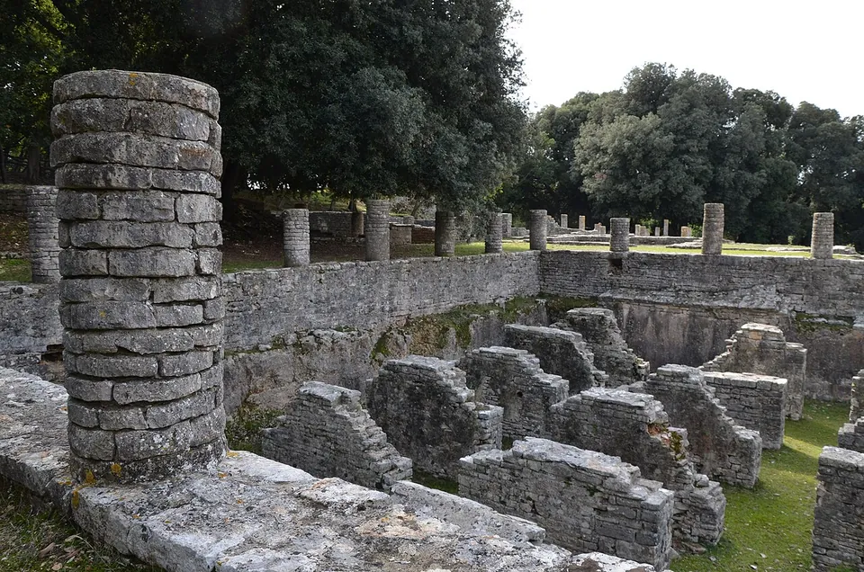 A column built of rough stone drums stands on a slab in the foreground; behind it a sunken area is divided by short stone walls into small compartments, below a terrace wall topped by a row of column stumps.