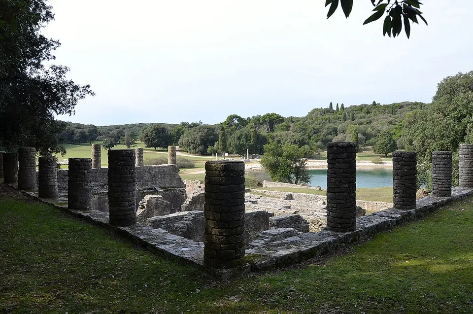 Short dark column shafts, some broken off, stand at intervals on a low stone foundation around a lawn, with excavated walls beyond and the bay visible between the trees.