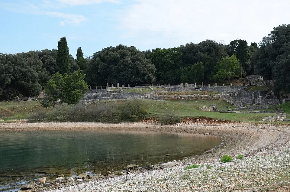 A calm bay with a pebbly shore curving round the foreground; across the water, pale stone walls and a row of short columns stand on a grassy terrace in front of dense trees.