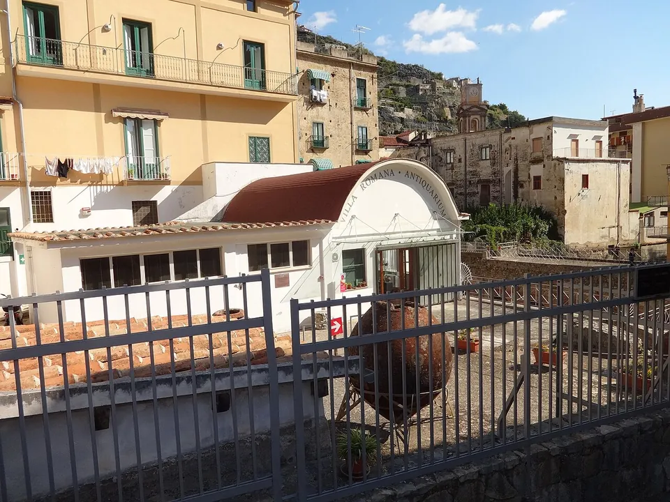 A low white building with a barrel-roofed porch lettered "Villa Romana · Antiquarium", a large terracotta jar on a metal stand in the paved yard in front, seen through grey railings, with tall houses and a hillside behind.