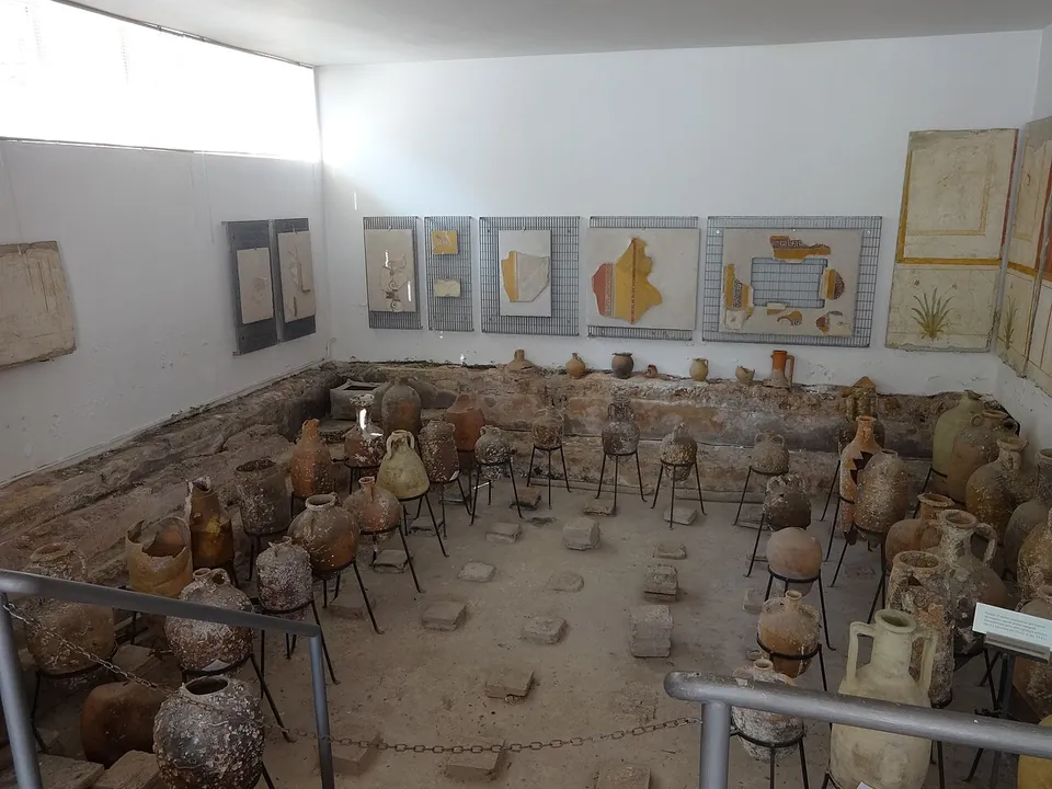 A white room with framed fragments of painted plaster on the walls and many amphorae and jars on metal stands across a sunken floor dotted with small square blocks.