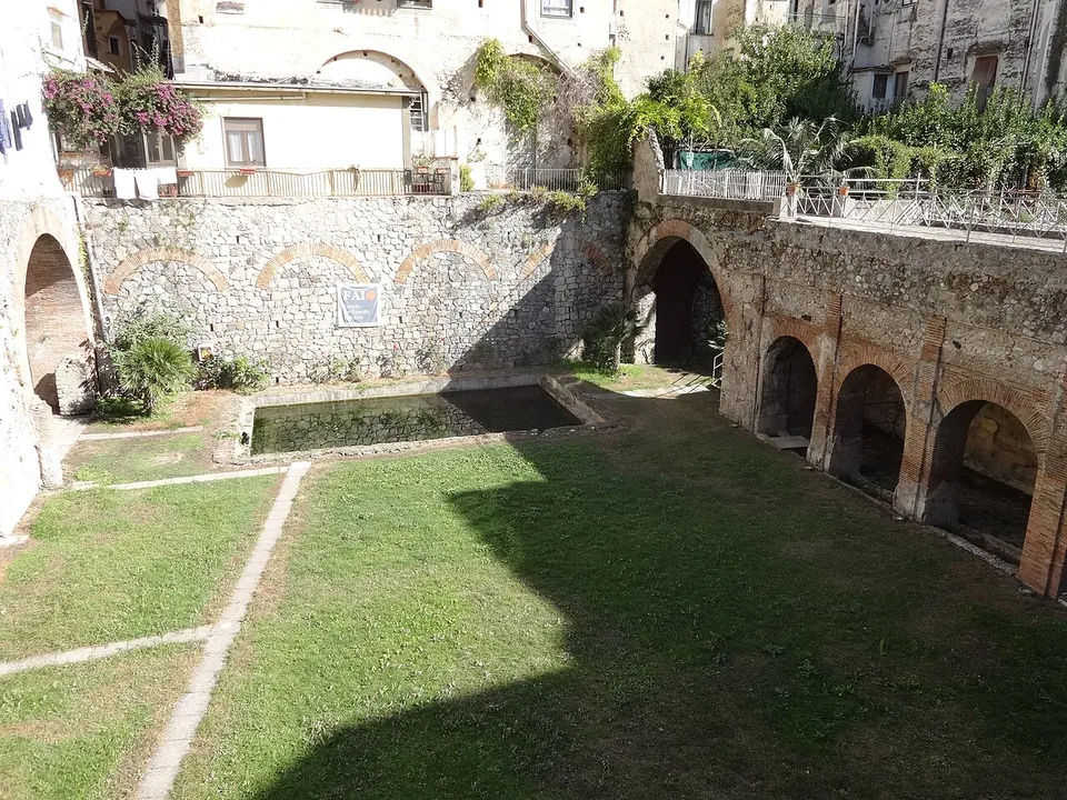 A sunken lawn with a long rectangular pool along one side, bounded by a row of round-headed arches in stone and brick, with the plastered houses of the modern town rising above the far wall.