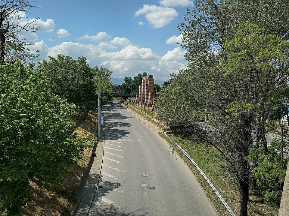 A tree-lined road seen from above, with the brick piers of the aqueduct standing in a grassy strip beside the carriageway in the middle distance and mountains on the horizon.