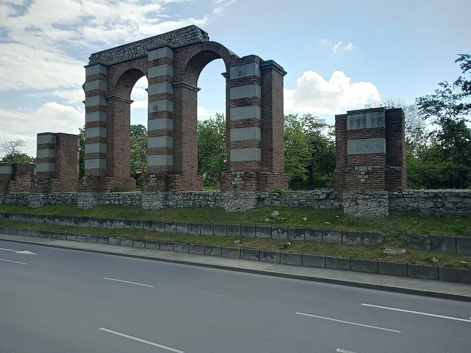 A row of tall piers of red brick banded with pale stone, three of them still joined by round arches, standing on a low stone wall along a grassy verge beside a road, under a blue sky with clouds.