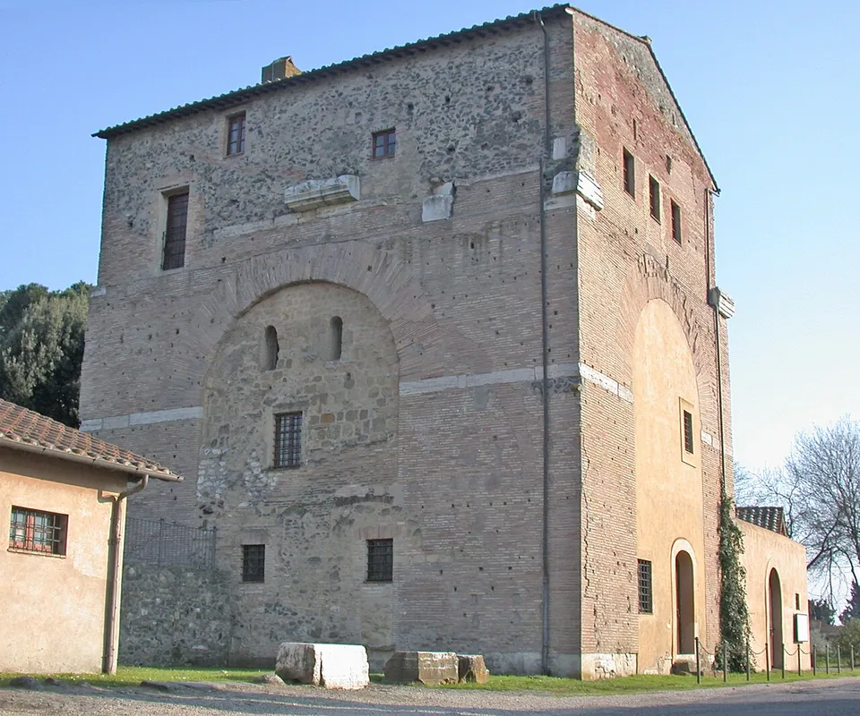 A tall, square building of brick and rubble with small windows and a tiled roof; the outline of a great round-headed arch, filled in with masonry, shows in the middle of the wall, and blocks of pale stone lie on the grass in front.