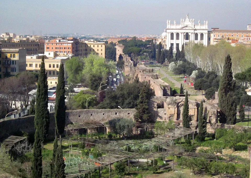 A view from high up over a walled garden of vegetable beds and wooden pergolas with tall cypresses, a long ruined wall running away to the right, and the white façade of a large church on the skyline beyond the rooftops.