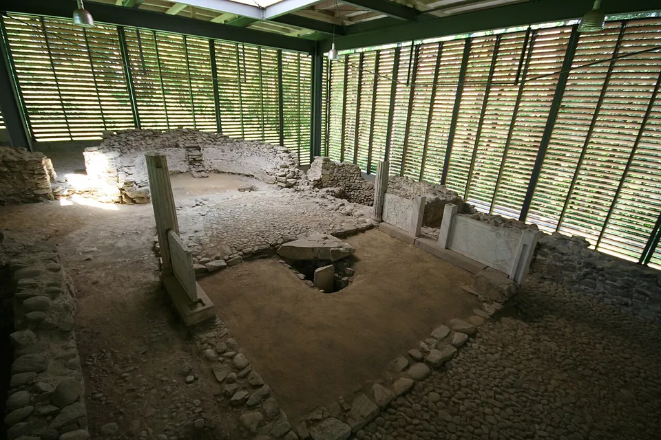 Low walls of rubble stone and a few upright stone posts on an earth floor, inside a large shelter of green steel and timber louvres.