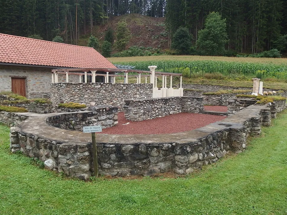Low rubble walls of a church laid out on grass, one rounded end filled with red gravel, beside a white building with a red tiled roof and a short row of white columns.