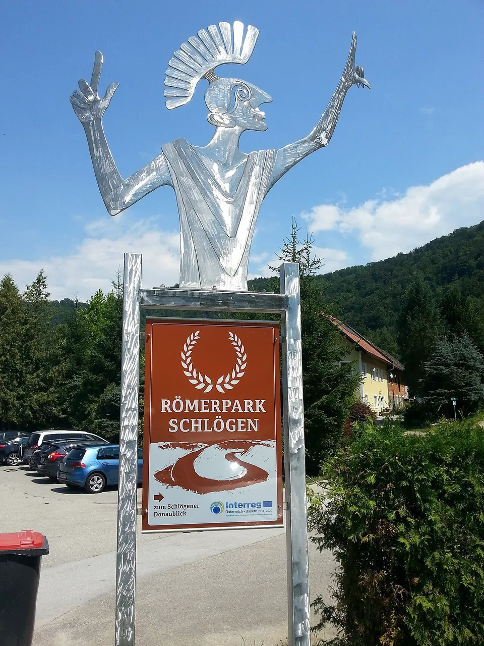 A brown sign reading "Römerpark Schlögen" on a metal post, topped by a stylised silver figure of a Roman soldier with a crested helmet, with a car park and wooded hills behind.