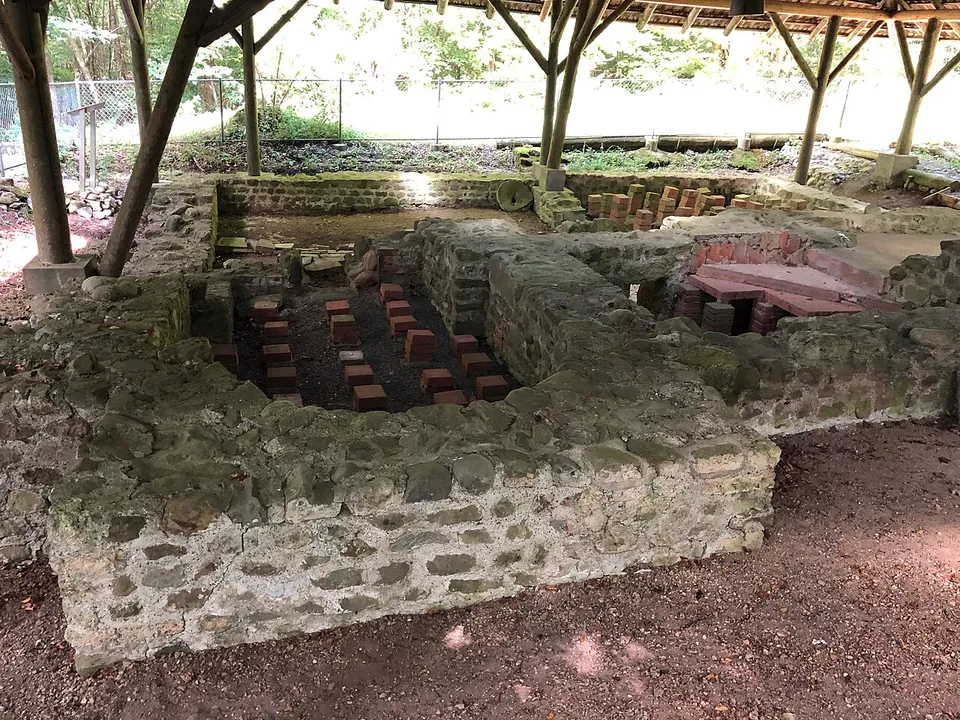 Low walls of rubble stone forming small rooms under an open timber roof, with rows of short square brick pillars standing in one room and tile slabs laid over pillars in another.
