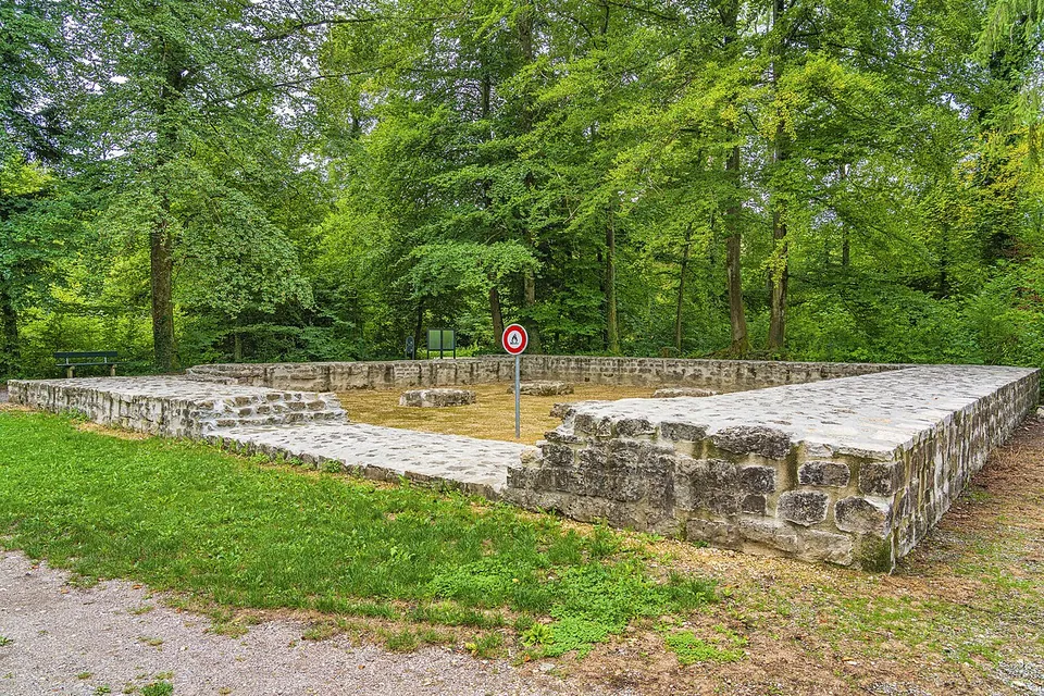 A square of low, neatly conserved stone walls on a gravel floor at the edge of a wood, with a lawn and a path in front.