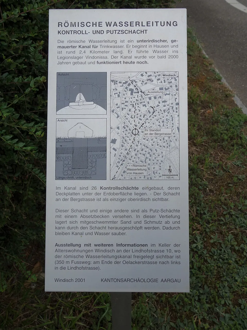 A white information board on a post in front of a hedge, headed "Römische Wasserleitung, Kontroll- und Putzschacht", with a cross-section drawing of the channel, a town map and German text.