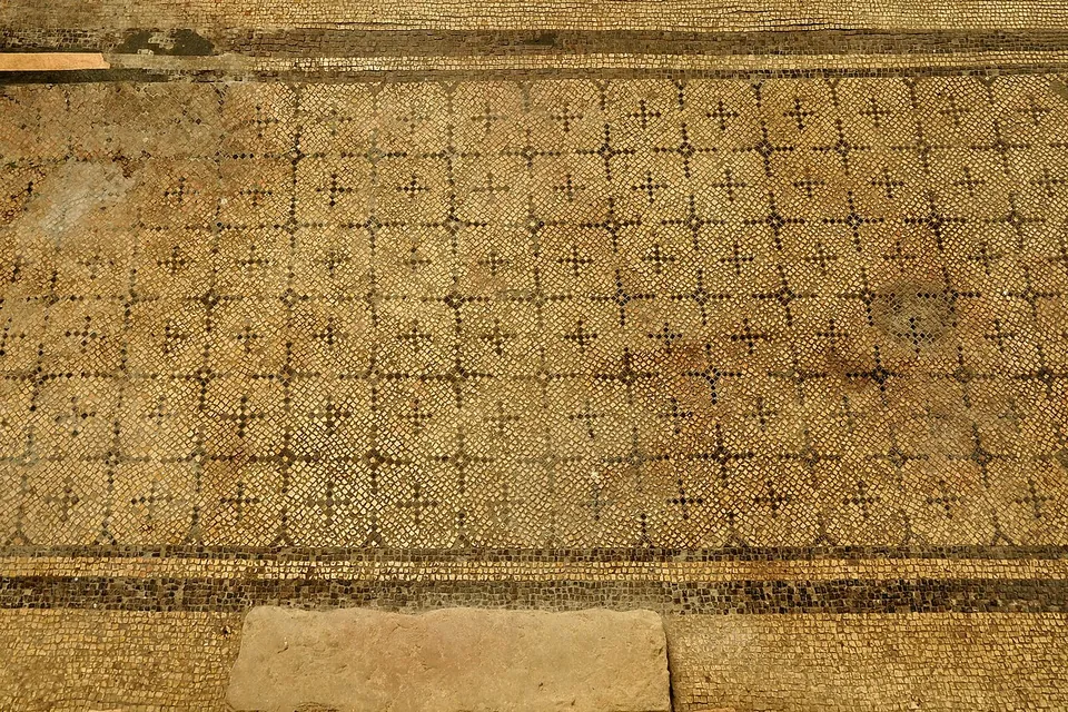 A worn mosaic floor of small light tesserae with a repeating grid of dark crosslets, edged by a dark border.