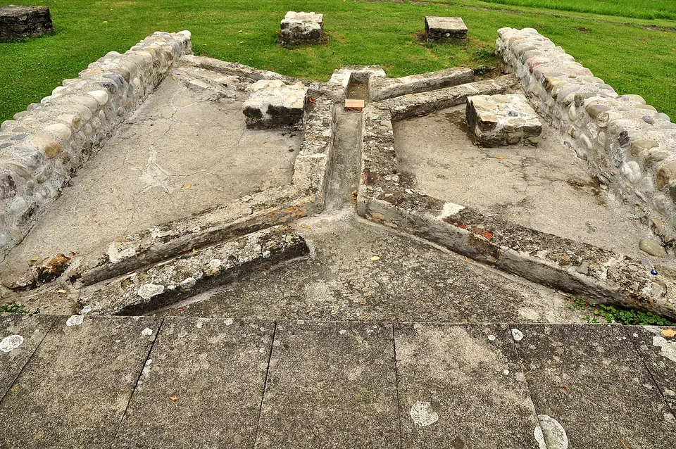 The low stone walls of a small room around a floor of grey mortar, crossed by an X of narrow channels edged with stones and brick, with grass and loose stone blocks beyond.