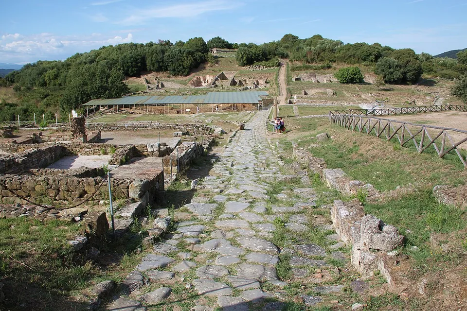 A Roman street paved with large irregular stone slabs runs up through the excavated town centre, with low stone walls of buildings on both sides, a long canopy roof over an excavation in the middle distance and wooded hills behind.