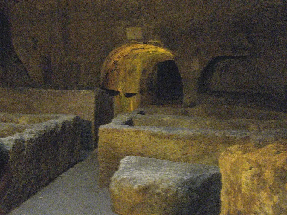 A dim rock-cut chamber whose floor is filled with rectangular stone tomb chests, with arched openings in the walls behind them.