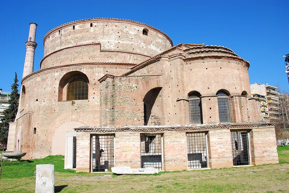 A massive round building of brick and stone under a low drum, with a later projecting wing with arched windows in front of it, a slender minaret on the left and grass with a few stone fragments in the foreground.
