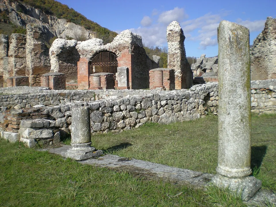Two weathered stone columns, one tall and one short, among low walls of rough stone in the grass, and behind them a row of ruined brick piers and arches, one closed with an iron grille, under a blue sky.