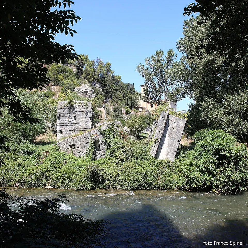 Across a fast-flowing river, two broken masses of squared stone masonry, one upright and one tilted, stand among bushes and trees on the far bank.
