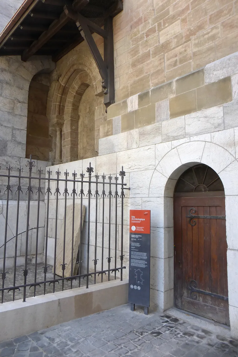 A small arched wooden door in the pale stone wall of the cathedral, beside an iron railing, with a red and grey sign on a post reading "Site Archéologique".