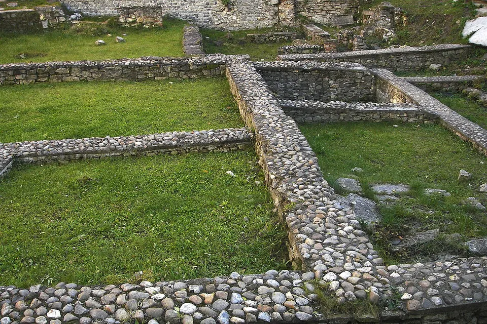 Low walls of rounded cobbles set in mortar outline several rooms in a mown lawn, with more ruined walls behind.