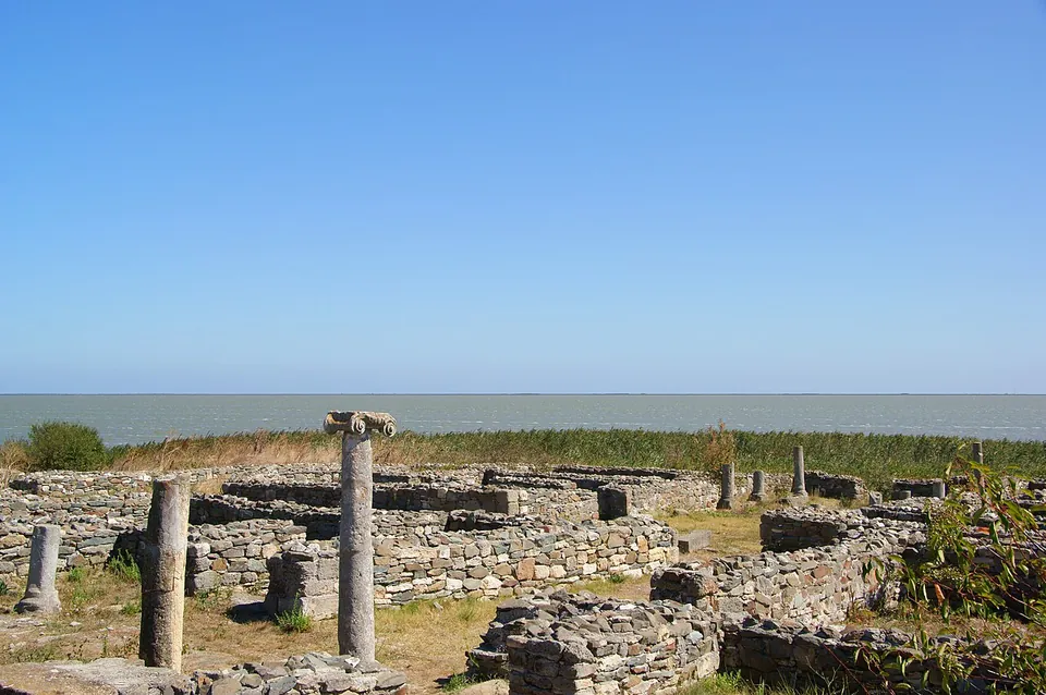 Several stone columns, one still with its capital, standing among low ruined walls on flat ground, with reeds and the wide water of the lake on the horizon.