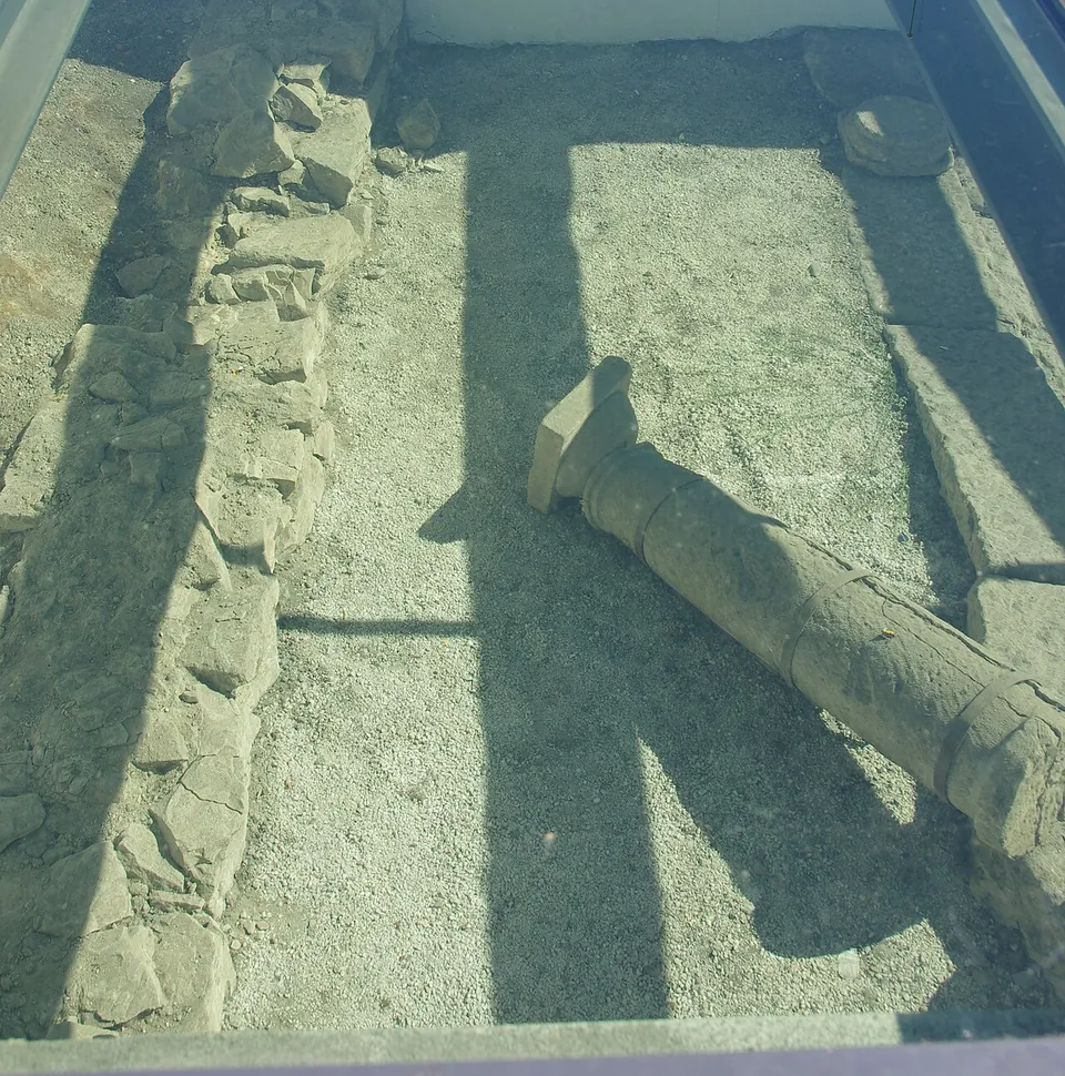 Looking down through a glass panel at a gravel floor, with a line of rough stone wall along one side and a stone column lying on its side, in strong sunlight.
