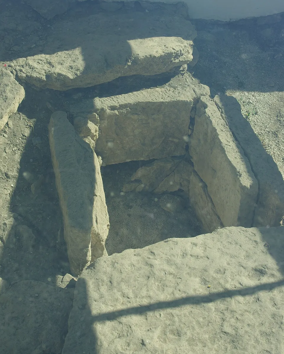 Close view through glass of large flat stone slabs set around a small square opening lined with upright stones.