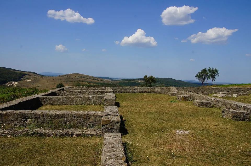 Low walls of excavated buildings laid out across a grassy hilltop, with rolling hills and a few trees on the horizon under a blue sky with small clouds.