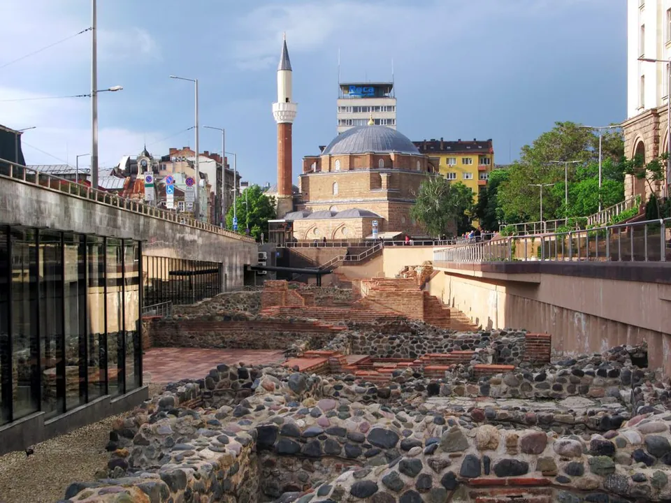 Low walls of rounded river stones and red brick in a sunken excavation in the city centre, with a mosque's dome and slender minaret behind it, modern office blocks and street lamps around it, under a grey sky.